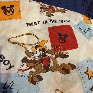 Puede incluir: Una tela de color claro con un dise&ntilde;o de Mickey Mouse con tem&aacute;tica de vaquero. Mickey est&aacute; montado a caballo y sosteniendo un lazo. Tambi&eacute;n se ve el texto "BEST IN THE WEST" y "Lil Buckaroo". El dise&ntilde;o incluye acentos naranjas, rojos y azules.