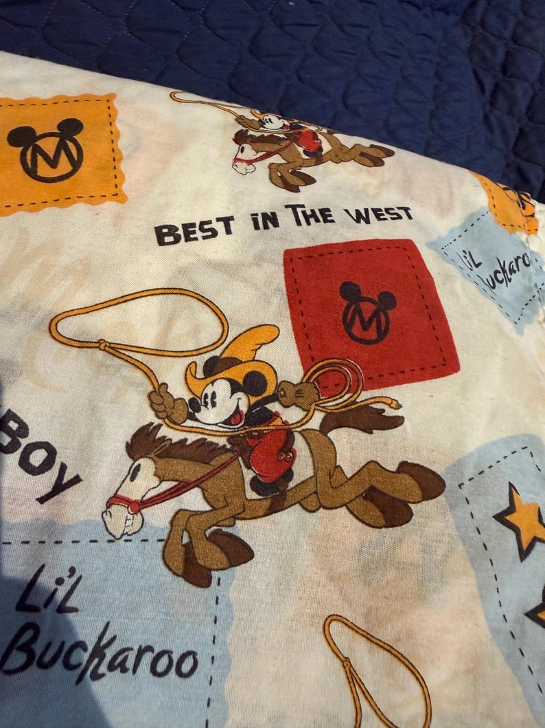 Puede incluir: Una tela de color claro con un dise&ntilde;o de Mickey Mouse con tem&aacute;tica de vaquero. Mickey est&aacute; montado a caballo y sosteniendo un lazo. Tambi&eacute;n se ve el texto "BEST IN THE WEST" y "Lil Buckaroo". El dise&ntilde;o incluye acentos naranjas, rojos y azules.