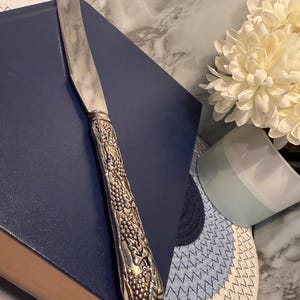 Puede incluir: Cuchillo de mantequilla plateado con un mango detallado con un diseño de uvas. El cuchillo está sobre un libro azul. La hoja es de acero inoxidable. El mango es plateado. Es un artículo vintage.