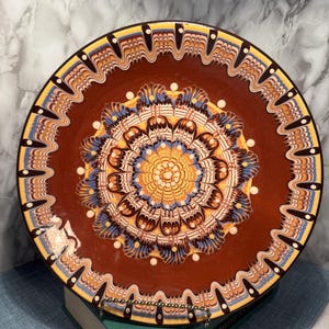 May include: A decorative ceramic plate with a brown base and intricate patterns in yellow, blue, and white. The plate features a central floral design and a detailed border, showcasing a traditional aesthetic.