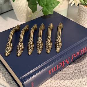 Op de afbeelding: Zes antieke messing kastgrepen in een tarwe-aren-ontwerp. De grepen zijn gerangschikt op een marineblauw boek met de rode woorden "Word Menu". De grepen zijn gemaakt van messing en hebben een gebogen vorm.