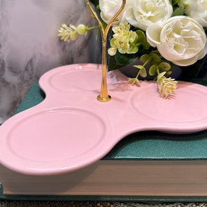May include: A pink, three-tiered serving tray with a gold handle. The tray has a unique, flowing shape with three circular sections. A bouquet of white flowers and greenery sits behind the tray, adding a touch of elegance.