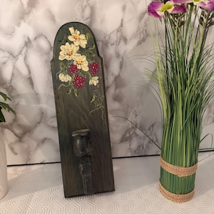 May include: A dark green wooden candle holder with a floral design. The candle holder has a curved top and a small cup to hold a candle. The flowers are painted in white and red. The candle holder is approximately 12 inches tall.