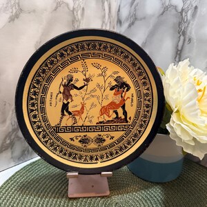 May include: A decorative plate with a black rim and gold center featuring a Greek mythological scene. The plate depicts figures, deer, and trees in black and orange against a gold background. The words "ARTEMIS" and "HRAKLIS" are written on the plate.