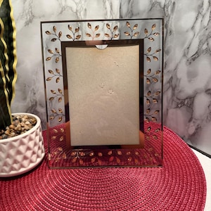 Pode incluir: Uma moldura retangular de vidro com borda dourada e detalhes de folhas. A moldura está vazia, com um fundo de cor neutra. A moldura é exibida em um tapete circular vermelho. Dimensões: 15 cm x 20 cm.