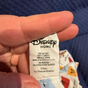 Puede incluir: Una etiqueta blanca de Disney Home se sostiene, mostrando el texto "TWIN FLAT SHEET" y "FINISHED SIZE 167 x 243 cm." La etiqueta tambi&eacute;n enumera la composici&oacute;n del material como 70% poli&eacute;ster y 30% algod&oacute;n. El logotipo de Disney es visible.