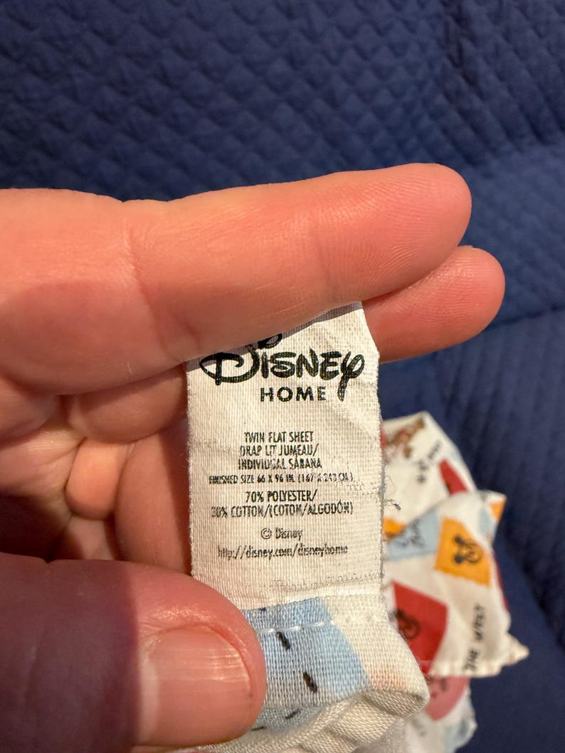 Puede incluir: Una etiqueta blanca de Disney Home se sostiene, mostrando el texto "TWIN FLAT SHEET" y "FINISHED SIZE 167 x 243 cm." La etiqueta tambi&eacute;n enumera la composici&oacute;n del material como 70% poli&eacute;ster y 30% algod&oacute;n. El logotipo de Disney es visible.