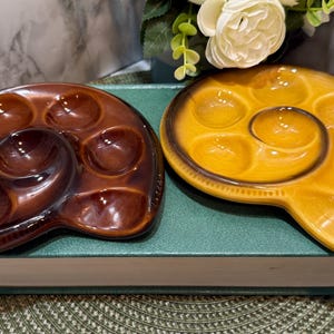 Könnte beinhalten: Zwei Schneckenplatten aus Keramik. Eine ist braun, die andere gelb mit braunem Rand. Jede Platte hat sechs kreisförmige Vertiefungen für die Schnecken und eine größere zentrale Vertiefung.