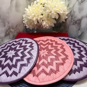 Puede incluir: Tres manteles individuales redondos con un diseño geométrico de estrella. Los manteles individuales son en tonos de rosa y morado. Los manteles individuales están sobre una superficie blanca con un fondo de mármol y un jarrón de flores blancas.