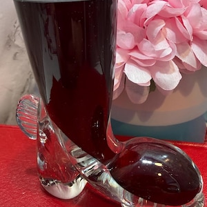 Puede incluir: Un jarrón decorativo en forma de bota de cristal burdeos. El jarrón se asienta sobre una base de cristal transparente. Se exhibe sobre una superficie roja, con un arreglo floral rosa en el fondo.