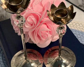 May include: Two silver candle holders with a flower-shaped top and a crystal accent. The candle holders are set against a backdrop of pink roses and a dark blue surface.
