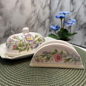 Puede incluir: Una mantequera de cerámica con tapa y un servilletero a juego. Ambas piezas están decoradas con motivos florales en tonos rosa, morado, amarillo y verde. El fondo presenta un patrón de mármol.
