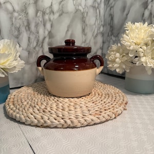 Puede incluir: Una olla de cerámica de dos tonos marrón y crema con tapa y dos asas. La olla está sobre un salvamanteles redondo tejido. Flores blancas en jarrones a ambos lados de la olla.