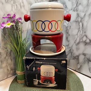 May include: A vintage fondue set with a white pot featuring colourful Olympic rings and red handles. The set includes a red stand, a white plate, and a black box with the product name. The set is designed for melting cheese or chocolate.