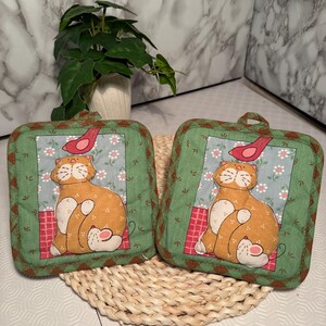 Puede incluir: Dos agarraderas cuadradas con un diseño de gato y pájaro. El gato naranja tiene detalles blancos, sentado sobre un patrón de cuadros rojos y blancos. El fondo es un estampado floral azul. El borde es verde con un lazo para colgar.