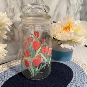 May include: A clear glass jar with a lid, decorated with red and green tulip illustrations. The jar has a cylindrical shape and is sitting on a blue and white braided mat. The word "Cutler" is printed on the jar.
