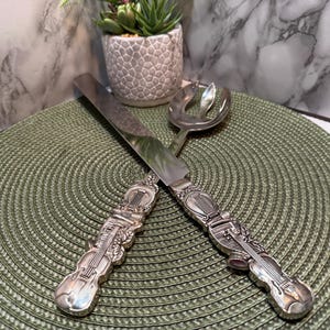 Könnte beinhalten: Silberfarbenes Servierbesteck-Set mit Tortenmesser und Servierlöffel. Die Griffe sind aufwendig mit Musikinstrumentenmotiven, darunter eine Geige und eine Harfe, gestaltet. Das Set liegt auf einem grünen Platzset.