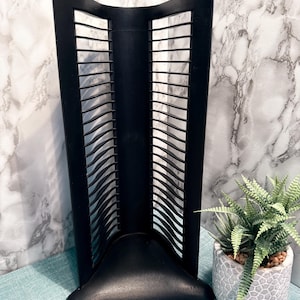 May include: Black CD or DVD storage rack with a triangular base and a series of horizontal dividers. The rack is made of a dark material and has a modern design. A small potted plant is next to the rack.