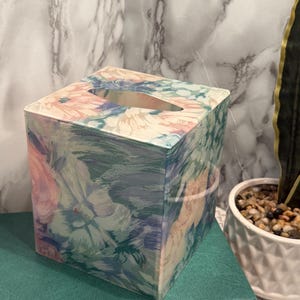 May include: A square tissue box with a floral design in pink, blue, and green. The box is placed on a green book. The tissue aperture is oval. The background includes a marble-effect wall and a potted plant.