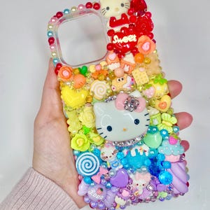 May include: A clear phone case decorated with colorful, three-dimensional embellishments. The design includes a Hello Kitty character, candies, flowers, and the word "Sweet". The case is adorned with beads and various charms.