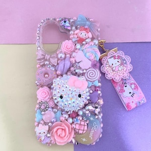 Puede incluir: Una funda de teléfono transparente decorada con una densa disposición de dijes de resina rosas, morados y azules, que incluyen una cara de Hello Kitty cubierta de pedrería, rosas, lazos, donas y paletas. Un dije de Hello Kitty a juego cuelga de una cinta.