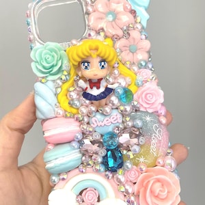 Peut inclure: Coque de téléphone transparente ornée d'une figurine Sailor Moon, de fleurs roses, de nœuds bleus et de décorations pastel. La coque présente un arc-en-ciel, des macarons et le mot "Sweet". Le design comprend des perles et des strass.