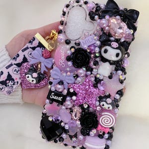 May include: A decorative phone case featuring a variety of embellishments. The case is adorned with black, pink, and purple beads, bows, and charms. The design includes a cartoon character, a star, and the word "Sweet". A matching wrist strap is attached.