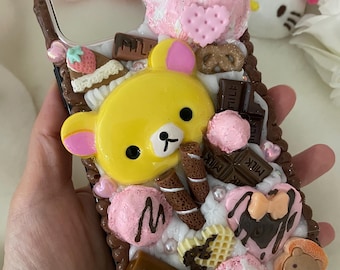 May include: A decorative phone case featuring a yellow bear character, pink ice cream, chocolate bars, and other dessert-themed embellishments. The case has a brown border and is adorned with various miniature food items.