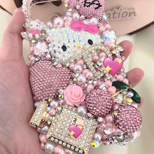 May include: A bedazzled pink phone case featuring a Hello Kitty design, embellished with rhinestones, pearls, and decorative elements. The case includes a pink bow, heart accents, and a perfume bottle design.