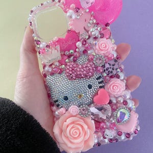 Può includere: Una custodia per telefono decorata con un design di Hello Kitty. La custodia è adornata con strass rosa e argento, perle ed elementi decorativi, tra cui un grande fiocco, rose e la scritta "Barbie".