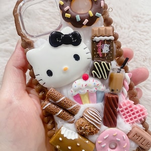 May include: A decorative phone case featuring a white Hello Kitty character with a black bow. The case is adorned with miniature sweets, including a chocolate donut, cupcake, and pink donut. Other details include chocolate bars and a drink.