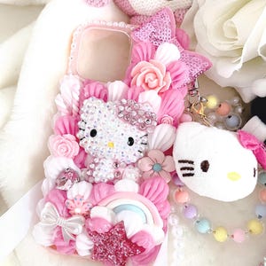 Peut inclure: Coque de téléphone rose ornée d'un motif Hello Kitty, entourée de fleurs roses et blanches, de nœuds et d'un arc-en-ciel. Un charm Hello Kitty en peluche est suspendu. La coque est ornée de strass et de paillettes.