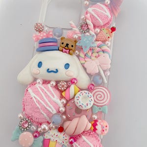 Peut inclure: Coque de téléphone transparente décorée d'une variété d'ornements colorés, fantaisistes et sur le thème des bonbons. Comprend un personnage de dessin animé blanc avec un chapeau bleu et rose, des bonbons roses, des beignets et un ours en peluche.