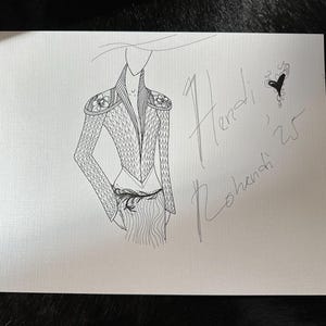 May include: A black and white fashion sketch of a jacket and skirt. The jacket has floral shoulder details and a textured pattern. The sketch includes the handwritten names "Hendi, Rohendi, 25" and a small heart.