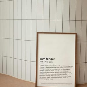 May include: A framed print with the text "sam fender" in a modern font. The print includes a brief biography of the British singer-songwriter known for anthemic indie rock. The frame is brown, and the background is a white tiled wall.