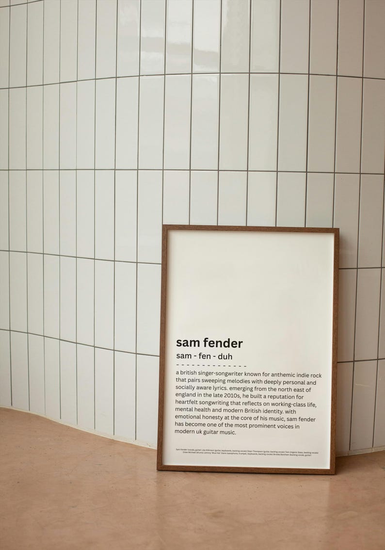 May include: A framed print with the text "sam fender" in a modern font. The print includes a brief biography of the British singer-songwriter known for anthemic indie rock. The frame is brown, and the background is a white tiled wall.