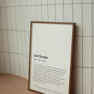 May include: A framed print with a dark brown wooden frame. The print features the name "sam fender" in a bold font, along with a brief biography of the British singer-songwriter. The background is white, and the frame is leaning against a white tiled wall.