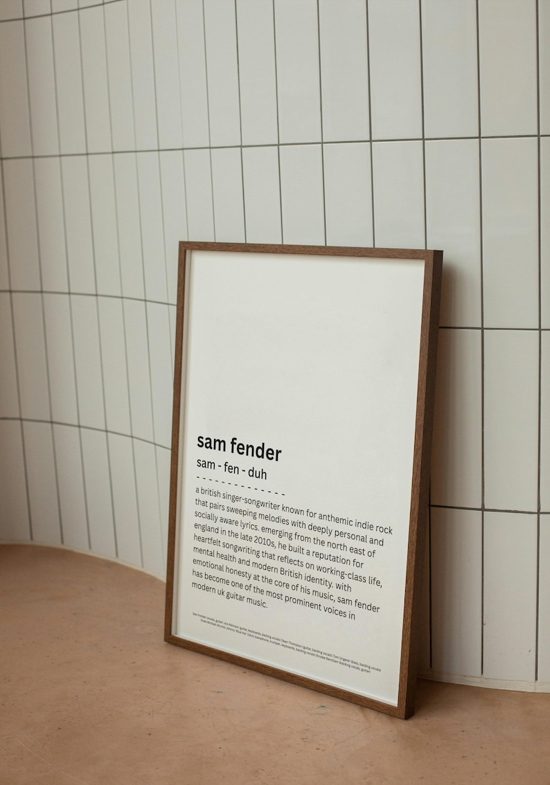 May include: A framed print with a dark brown wooden frame. The print features the name "sam fender" in a bold font, along with a brief biography of the British singer-songwriter. The background is white, and the frame is leaning against a white tiled wall.
