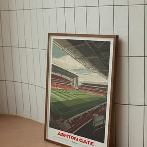 Op de afbeelding: Een ingelijste print van het Ashton Gate stadion in Bristol City. Het kunstwerk toont het interieur van het stadion met een groen veld, rode stoelen en een gedeeltelijke weergave van de lucht. De print zit in een bruine houten lijst.