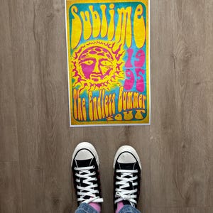 May include: A poster with the text "Sublime 1995 The Endless Summer Tour" in yellow, pink, and blue. The poster features a sun graphic with a face. Black Converse sneakers with white laces and soles are visible.