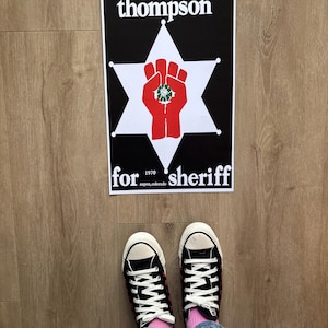 May include: A black and white poster with the text "thompson for sheriff" and a red fist inside a white star. The poster is on a wooden floor, with black Converse trainers and pink socks visible.