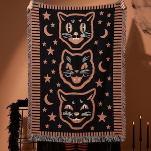 Peut inclure: Un plaid tissé rectangulaire à fond noir orné de trois illustrations superposées de têtes de chats noirs en orange et blanc. Le motif comprend des croissants de lune et des étoiles orange, avec une bordure rayée et des franges aux extrémités.