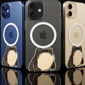 May include: Three smartphones in blue, gray, and gold, each encased in a clear protective cover. Each case features a white circular magnetic ring centered on the back and a graphic illustration of a frog with a light tan belly and dark outlines.