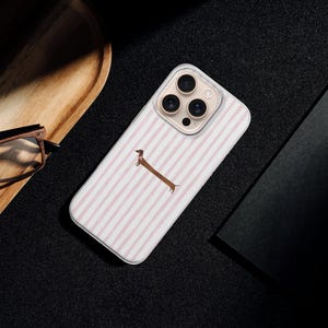 May include: A phone case with a white and pink striped design and a brown dachshund illustration. The case is on a black surface, alongside a wooden tray, glasses, and a black notebook.
