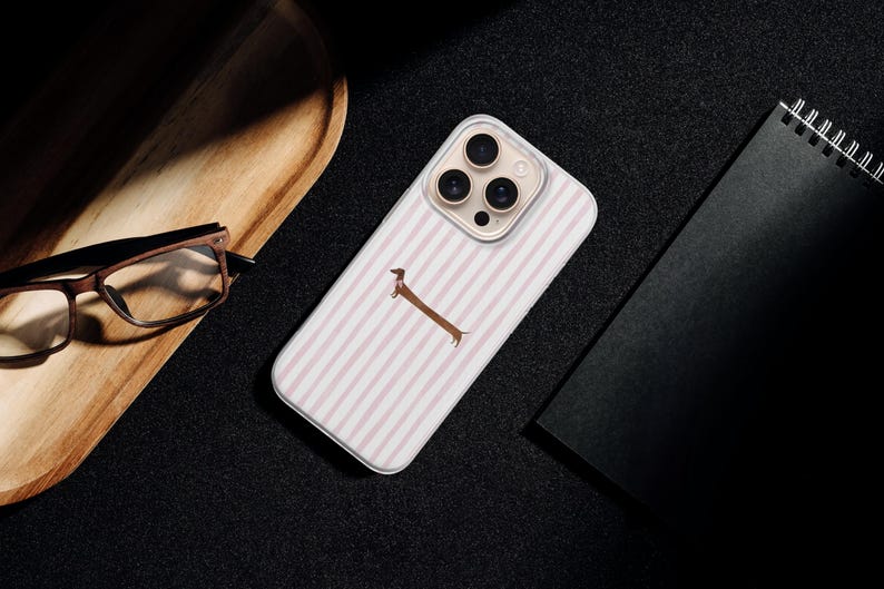 May include: A phone case with a white and pink striped design and a brown dachshund illustration. The case is on a black surface, alongside a wooden tray, glasses, and a black notebook.