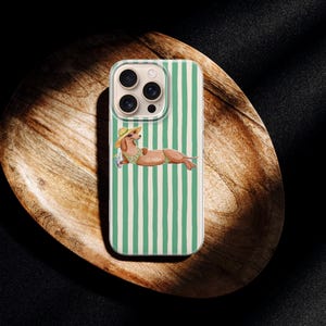 May include: A phone case with a green and white striped design. The case features a dachshund illustration wearing a hat and sunglasses, relaxing on a beach towel. The phone case is on a wooden tray.