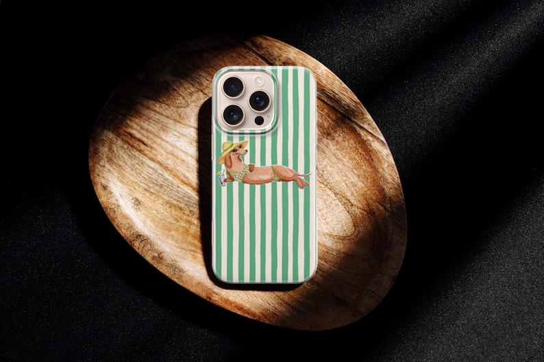 May include: A phone case with a green and white striped design. The case features a dachshund illustration wearing a hat and sunglasses, relaxing on a beach towel. The phone case is on a wooden tray.