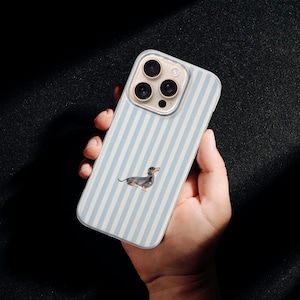 May include: A phone case with a light blue and white striped pattern, featuring a dachshund dog illustration. The case has a clear camera lens area. The phone case is held against a dark background.
