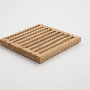 May include: A square wooden breadboard with parallel grooves. The board is made of light-coloured wood and has rounded edges. The grooves are designed to catch crumbs. A kitchen item.