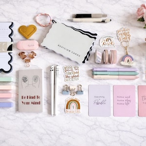 May include: An assortment of stationery items arranged on a surface. The image includes a daily planner, revision cards, pens, washi tape, and inspirational cards. The color scheme features pastels, gold, and black. A glass jar with a rainbow design and pink roses are also visible.